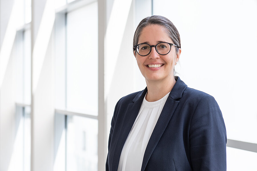 Frau mit dunklen Haaren und Brille in einem schwarzen Blazer im Portrait. Im Hintergrund ein Glasgang. Frau mit dunklen Haaren und Brille in einem schwarzen Blazer im Portrait. Im Hintergrund ein Glasgang.