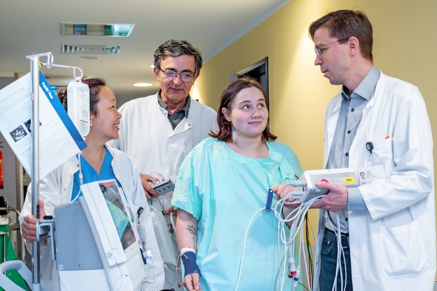 Drei Personen in medizinischer Arbeitskleidung stehen auf einem Klinikflur. Zwischen ihnen steht eine Patientin in Krankenhauskleidung. Die Person rechts im Bild hält ein medizinisches Gerät in den Händen. Auch links im Bild befindet sich ein medizinisches Gerät.  Drei Personen in medizinischer Arbeitskleidung stehen auf einem Klinikflur. Zwischen ihnen steht eine Patientin in Krankenhauskleidung. Die Person rechts im Bild hält ein medizinisches Gerät in den Händen. Auch links im Bild befindet sich ein medizinisches Gerät.