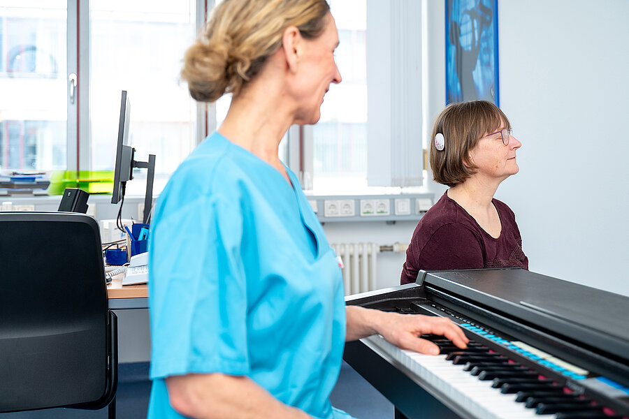Eine Patientin sitzt mit geschlossenen Augen in einem Behandlungsraum eines Krankenhauses. An ihrem Kopf befindet sich ein Cochlea-Implantat. Im Vordergrund sitzt eine Frau in medizinischer Arbeitskleidung an einem Klavier. Eine Patientin sitzt mit geschlossenen Augen in einem Behandlungsraum eines Krankenhauses. An ihrem Kopf befindet sich ein Cochlea-Implantat. Im Vordergrund sitzt eine Frau in medizinischer Arbeitskleidung an einem Klavier.