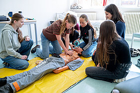 Eine Schülerin führt an einer Puppe eine Herzdruckmassage durch. Eine kleine Gruppe von Schülerinnen sitzt um die Puppe herum und schaut zu.