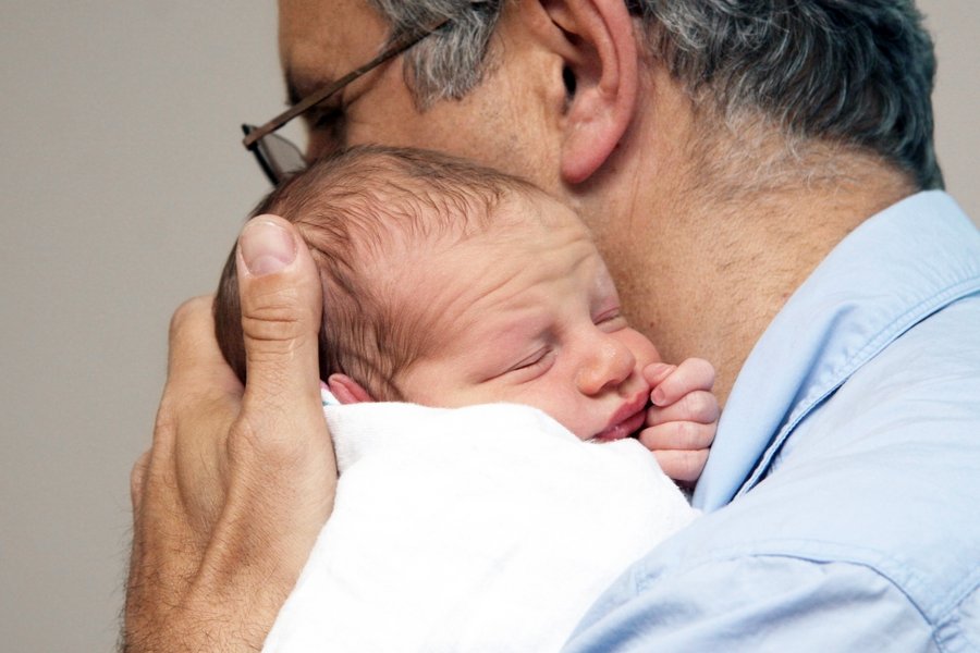 Ein älterer Mann hält ein neugeborenes Baby fest an sich gedrückt. Das Baby sieht noch etwas zerknautscht aus. Die Szene wirkt sehr liebevoll.    Ein älterer Mann hält ein neugeborenes Baby fest an sich gedrückt. Das Baby sieht noch etwas zerknautscht aus. Die Szene wirkt sehr liebevoll.  