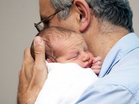 Ein älterer Mann hält ein neugeborenes Baby fest an sich gedrückt. Das Baby sieht noch etwas zerknautscht aus. Die Szene wirkt sehr liebevoll.  