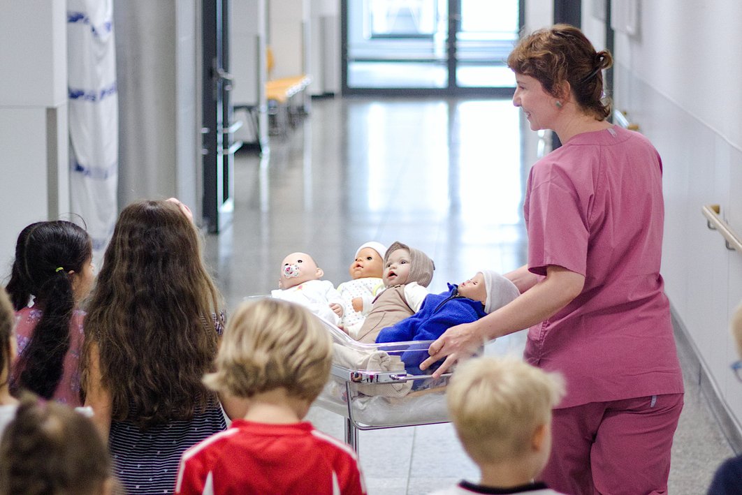 Eine Hebamme in beerenfarbener medizinischer Arbeitskleidung schiebt ein Krankenhaus-Babybettchen, in dem mehrere Babypuppen liegen, einen Gang entlang. Hinter ihr laufen mehrere Kinder im Vorschulalter. Blick von hinten auf die Gruppe.