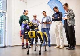 Eine Gruppe von fünf Personen steht hinter einem Roboter, der wie ein Hund gebaut ist. Der Roboter ist schwarz-gelb gefärbt und blickt an der Kamera vorbei. 