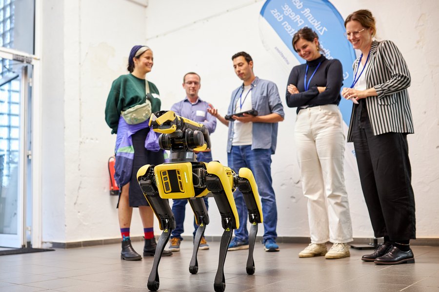 Eine Gruppe von fünf Personen steht hinter einem Roboter, der wie ein Hund gebaut ist. Der Roboter ist schwarz-gelb gefärbt und blickt an der Kamera vorbei.   Eine Gruppe von fünf Personen steht hinter einem Roboter, der wie ein Hund gebaut ist. Der Roboter ist schwarz-gelb gefärbt und blickt an der Kamera vorbei. 