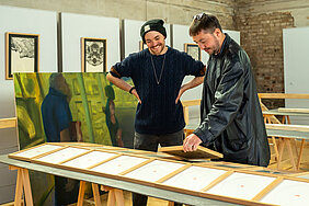 Zwei Personen stehen in einem alten Gemäuer, an dessen Wänden zahlreiche Bilderrahmen und Galeriewände lehnen. Die eine Person hat die Hände in die Hüften gestemmt und lacht, während sie ein Ausstellungsstück betrachtet. Die andere Person stellt das Exponat auf den dafür vorgesehenen Platz und lächelt.