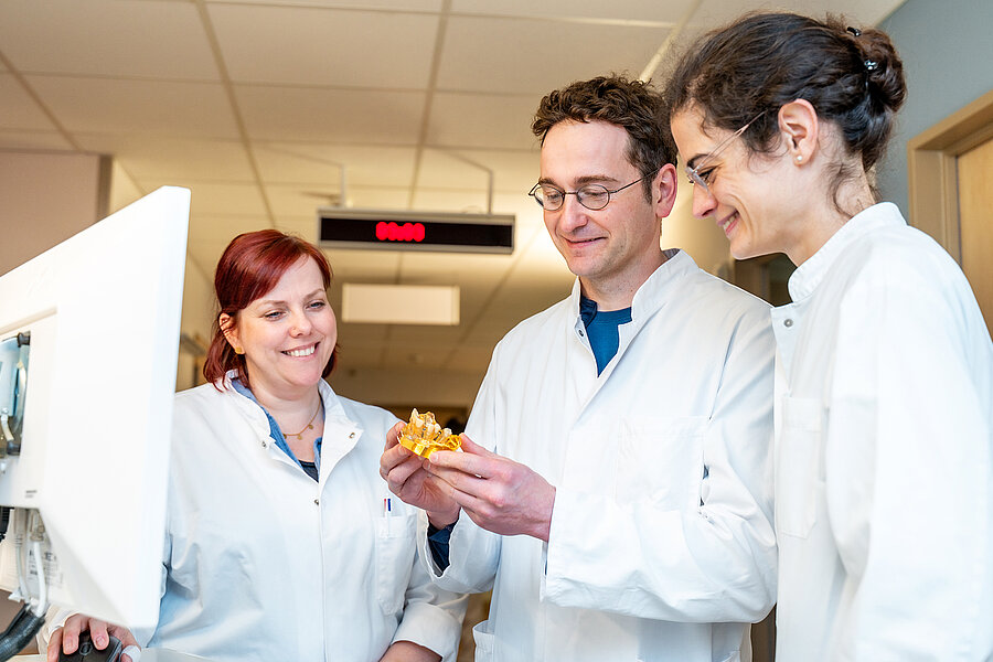 Drei Personen in medizinischer Arbeitskleidung stehen zusammen auf einem Krankenhausflur. Die Person in der Mitte hält ein kleines zahnmedizinisches Modell in der Hand. Drei Personen in medizinischer Arbeitskleidung stehen zusammen auf einem Krankenhausflur. Die Person in der Mitte hält ein kleines zahnmedizinisches Modell in der Hand.