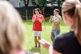 Eine Frau und ein Mann stehen auf einer Wiese und begrüßen die Teilnehmenden eines Sportfests. Die Frau lächelt, beide Personen klatschen in die Hände. Die Frau trägt sportliche Kleidung, der Mann eine Jeans, ein Hemd und einen Pullover. Im Vordergrund sind zwei Personen unscharf von hinten abgebildet. 