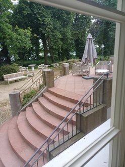 Vom großen Salon aus geht es auf eine Terrasse und führt eine Freitreppe in den parkähnlichen Garten mit Sitzgelegenheiten. Die Steintreppe ist rötlich, flankiert von Metallgeländern, auf der Terrasse stehen graue, geschlossene Sonnenschirme. Getrennt durch einen breiten Kiesweg schließt sich eine Grünfläche mit großen alten Bäumen an, unter denen unter anderem Holzstühle stehen.