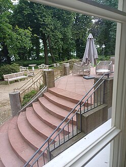 Vom großen Salon aus geht es auf eine Terrasse und führt eine Freitreppe in den parkähnlichen Garten mit Sitzgelegenheiten. Die Steintreppe ist rötlich, flankiert von Metallgeländern, auf der Terrasse stehen graue, geschlossene Sonnenschirme. Getrennt durch einen breiten Kiesweg schließt sich eine Grünfläche mit großen alten Bäumen an, unter denen unter anderem Holzstühle stehen.