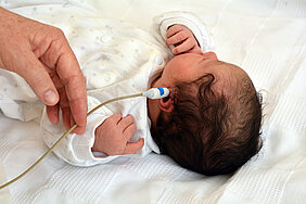 Ein Baby liegt auf einem Bett oder einer Liege. In seinem linken Ohr befindet sich ein kleines medizinisches Gerät für das Neugeborenen-Hörscreening. Eine Hand einer Person hält das Kabel, das zu dem Gerät gehört.