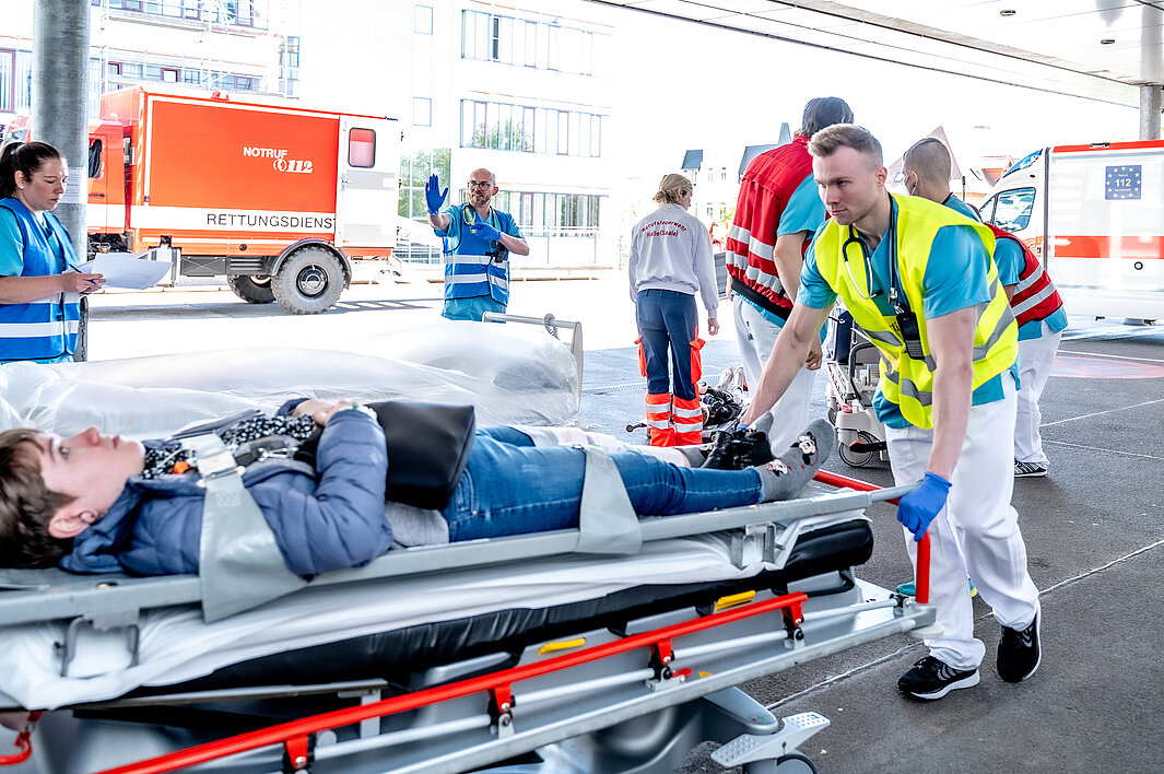 Eine Person in medizinischer Arbeitskleidung schiebt eine Person auf einer Trage in die Notaufnahme. Im Hintergrund befinden sich weitere Personen, die an der Katastrophenschutzübung mitwirken.