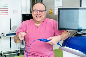 Eine Person in medizinischer Arbeitskleidung befindet sich in einem Raum eines Klinikums und schaut in die Kamera. Die Person hält ein medizinisches Gerät in den Händen. Rechts neben der Person befindet sich ein Monitor.