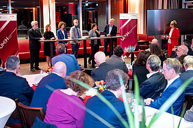 Viele Menschen sitzen an Tischen. Im Bildhintergrund sind mehrere Teilnehmende einer Podiumsdiskussion zu sehen.