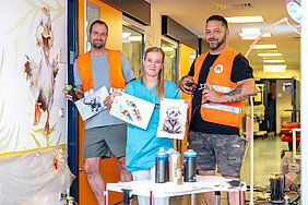 Drei Personen stehen im Flur einer Kinderstation in einem Krankenhaus. Die Person in der Mitte trägt medizinische Arbeitskleidung und hält Entwürfe für die Wandgestaltung der Station in den Händen. Die beiden Personen außen tragen orange Warnwesten und halten Gestaltungsutensilien und einen Entwurf in den Händen. Auf einem Tisch im Vordergrund stehen Sprühdosen. Links im Bild befindet sich eine Ente als Teil der Wandgestaltung.