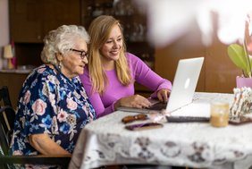 Eine ältere und eine jüngere Frau sitzen im häuslichen Umfeld vor einem Laptop an einem Tisch. Auf der Tischdecke mit Spitze liegen weitere Alltagsgegenstände wie Sonnenbrille, Handy und Fernbedienung. Die beiden Frauen lächeln und blicken auf den Laptop. Im Hintergrund ist unscharf eine große dunkelbraune Anbauwand zu erkennen.