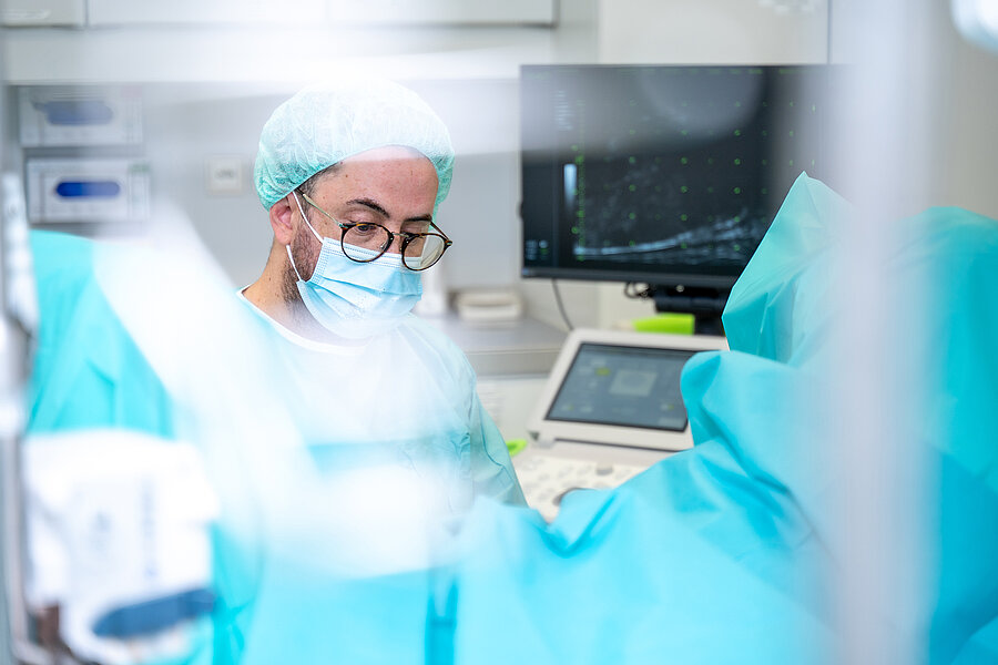 Bildvordergrund in blauer Unschärfe. Im Hintergrund ist ein Mann mit medizinischer Schutzkleidung und ein Ultraschallmonitor zu sehen. Bildvordergrund in blauer Unschärfe. Im Hintergrund ist ein Mann mit medizinischer Schutzkleidung und ein Ultraschallmonitor zu sehen.