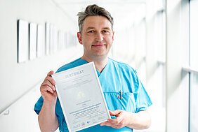 Eine Person in medizinischer Arbeitskleidung steht in einem Krankenhausflur und hält ein Zertifikat in den Händen.