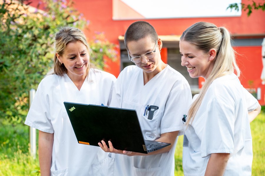 Drei Personen stehen nebeneinander auf einer Wiese. Eine Person hält einen Laptop in den Händen und zeigt den anderen Personen etwas auf dem Bildschirm. Im Hintergrund befinden sich ein Strauch und ein Gebäude.  Drei Personen stehen nebeneinander auf einer Wiese. Eine Person hält einen Laptop in den Händen und zeigt den anderen Personen etwas auf dem Bildschirm. Im Hintergrund befinden sich ein Strauch und ein Gebäude.