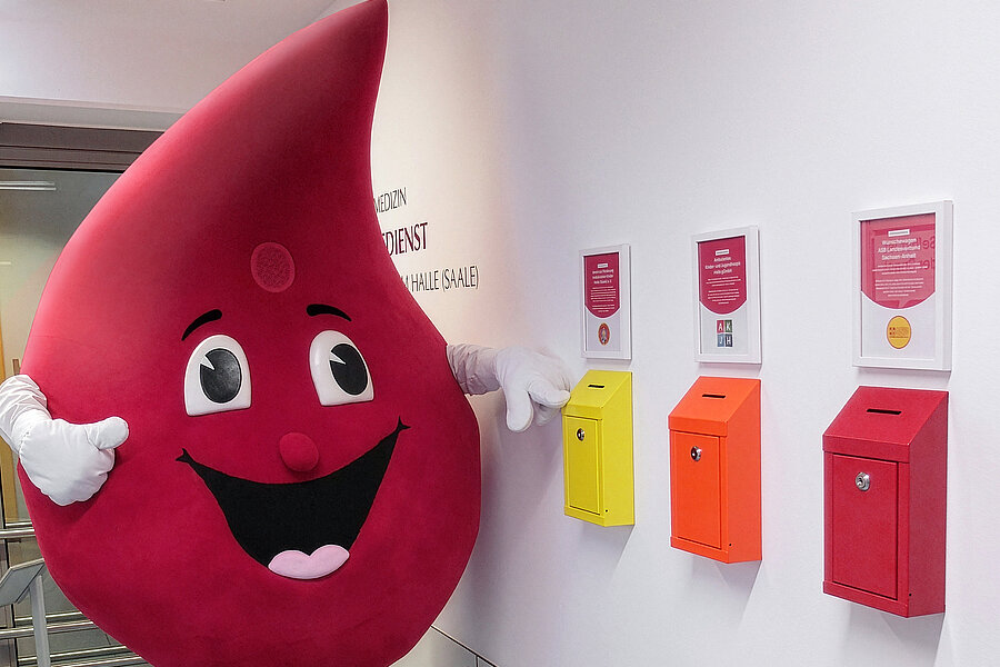 Das Maskottchen "Bob the Drop" der Blutspende des Universitätsklinikums Halle (Saale), ein großer Blutstropfen, steht neben drei Spendenboxen, die an einer Wand befestigt sind. Das Maskottchen "Bob the Drop" der Blutspende des Universitätsklinikums Halle (Saale), ein großer Blutstropfen, steht neben drei Spendenboxen, die an einer Wand befestigt sind.