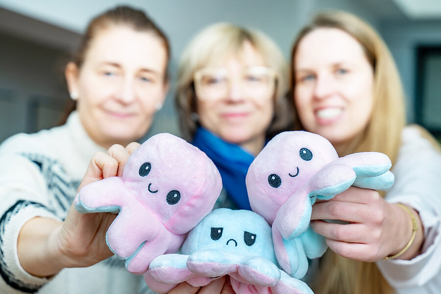 Drei Frauen halten lächelnde Stoff-Kraken in die Kamera; die Figuren sind rosa und blau und dienen als Maskottchen eines Angebots der Psychiatrischen Institutsambulanz für Kinder. Drei Frauen halten lächelnde Stoff-Kraken in die Kamera; die Figuren sind rosa und blau und dienen als Maskottchen eines Angebots der Psychiatrischen Institutsambulanz für Kinder.