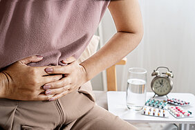 Detailaufnahme einer Person von der Brust bis zu den Oberschenkeln. Die Person hält sich mit beiden Händen den Bauch. Rechts neben ihr befinden sich ein Wecker, ein Wasserglas und Medikamente auf einer Ablage.