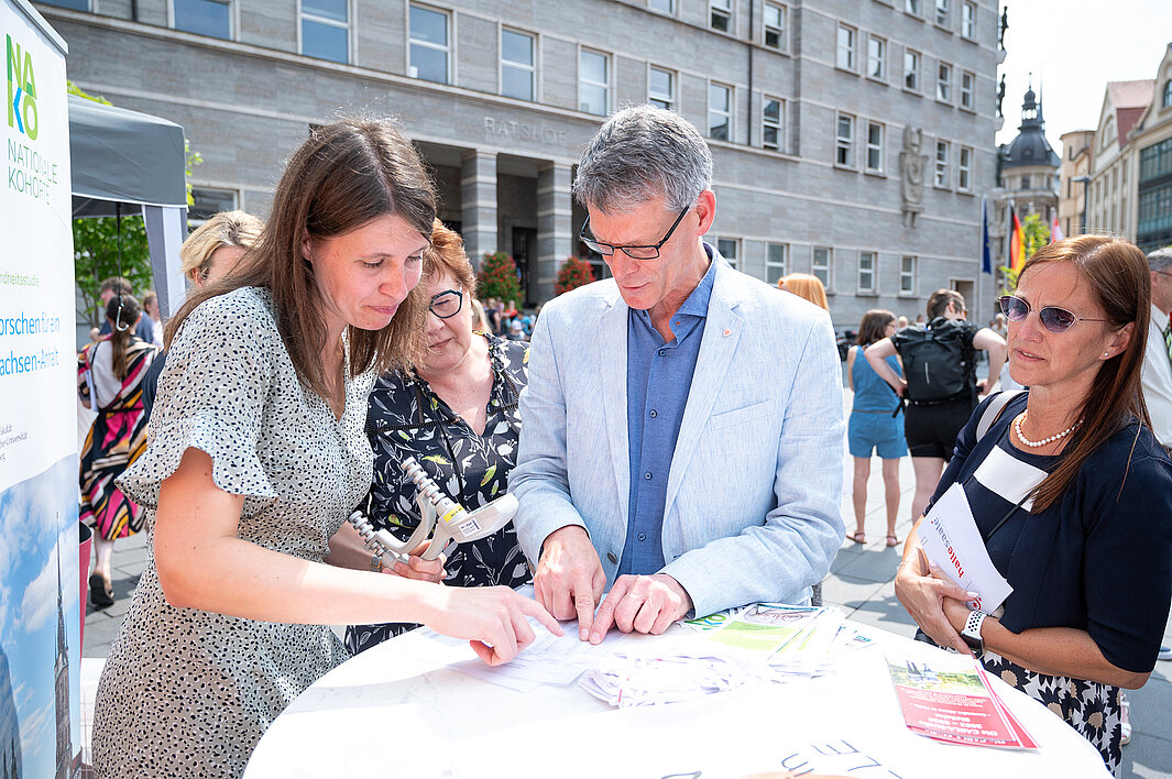 Bürgermeister Egbert Geier steht zwischen zwei Frauen an einem Stehtisch. Eine Frau erklärt etwas anhand eines Flyers.