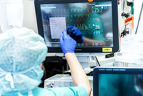 Eine Person, die eine OB-Haube und medizinische Einmalhandschuhe trägt, bedient einen Bildschirm. Auf dem Display sind verschiedene Vitalparameter wie Puls, Blutdruck und Sauerstoffsättigung erkennbar.