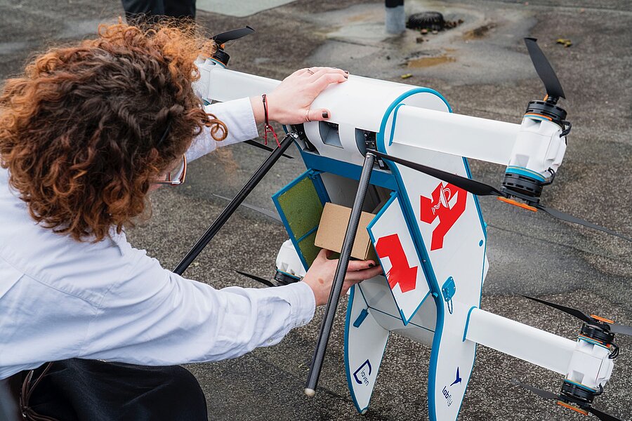 Eine Frau verstaut einen kleinen Pappkarton in einer Drohne. Die Drohne ist etwas größer als ein Umzugskarton, hat acht handgroße Rotoren und ist auf der Seite mit einem knallroten Apothekensymbol versehen. Eine Frau verstaut einen kleinen Pappkarton in einer Drohne. Die Drohne ist etwas größer als ein Umzugskarton, hat acht handgroße Rotoren und ist auf der Seite mit einem knallroten Apothekensymbol versehen.