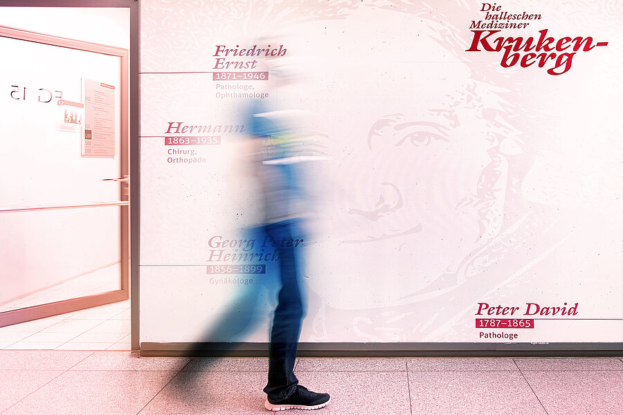 Eine Person läuft einen Flur entlang. Die Person befindet sich in der Bewegung und ist nur schemenhaft dargestellt. Im Hintergrund befindet sich eine Wand mit dem Schriftzug "Die halleschen Mediziner Krukenberg" sowie weiteren Namen und Daten. Eine Person läuft einen Flur entlang. Die Person befindet sich in der Bewegung und ist nur schemenhaft dargestellt. Im Hintergrund befindet sich eine Wand mit dem Schriftzug "Die halleschen Mediziner Krukenberg" sowie weiteren Namen und Daten.