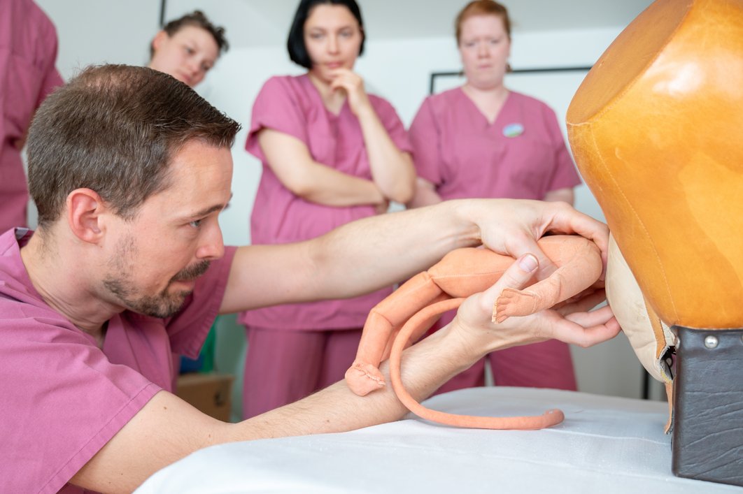 Ein Arzt zieht eine Babypuppe aus einem Geburtsmodell aus Holz. Er schaut sehr konzentriert. Im Hintergrund sieht man einige medizinische Mitarbeiterinnen, die aufmerksam zuschauen. Alle tragen beerenfarbene medizinische Arbeitskleidung.