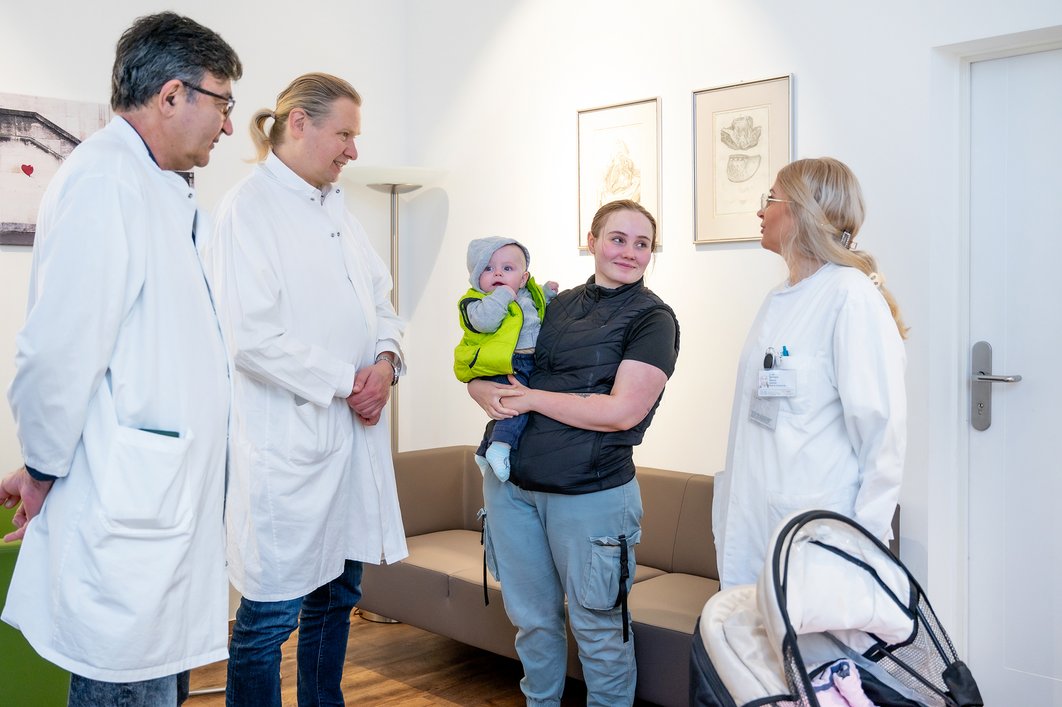 Drei Personen in medizinischer Berufskleidung stehen in einem Büro. Zwischen ihnen steht eine Frau, die ein Baby auf dem Arm trägt.