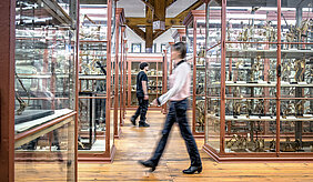 Ein großer Raum mit zahlreichen Vitrinen. Im Mittelgang sind zwei Personen in Bewegungsunschärfe erkennbar, die zwischen den Vitrinen hin und her laufen. In den Vitrinen sind Skelette verschiedener Arten ausgestellt, darunter Vögel und Raubtiere. Außerdem sind die historischen, hohen Decken und die restaurierte Dachbalkenkonstruktion gut zu erkennen.