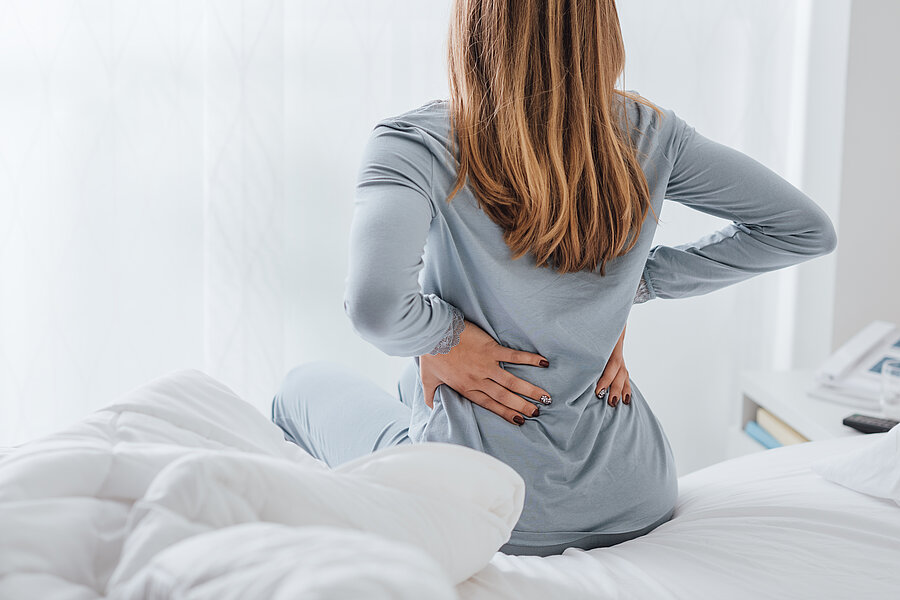 Frau von hinten auf einem Bett sitzend. Sie fasst sich mit beiden Henden an den unteren Rücken. Frau von hinten auf einem Bett sitzend. Sie fasst sich mit beiden Henden an den unteren Rücken.