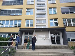 Außenansicht des 2. HMU-Standortes, der ein ehemaliges DDR-typisches Schulgebäude ist. Die Fassade ist gelb gestrichen, zum Eingang führt eine breite, mehrstufige Treppe. Links ist zudem eine Rampe vorhanden.