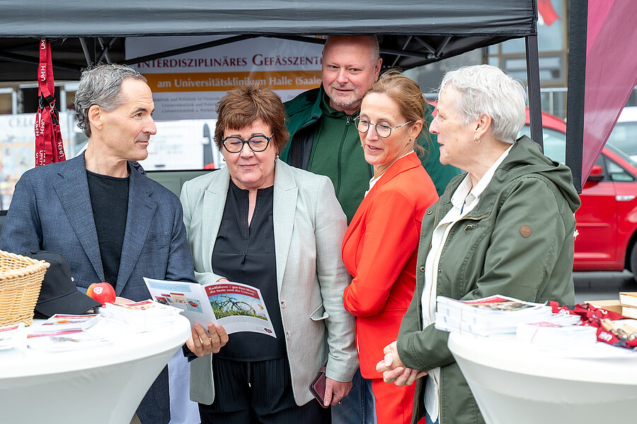 Fünf Personen stehen an einem Infostand an zwei Stehtischen. Eine Person hält einen Flyer in der Hand. Fünf Personen stehen an einem Infostand an zwei Stehtischen. Eine Person hält einen Flyer in der Hand.