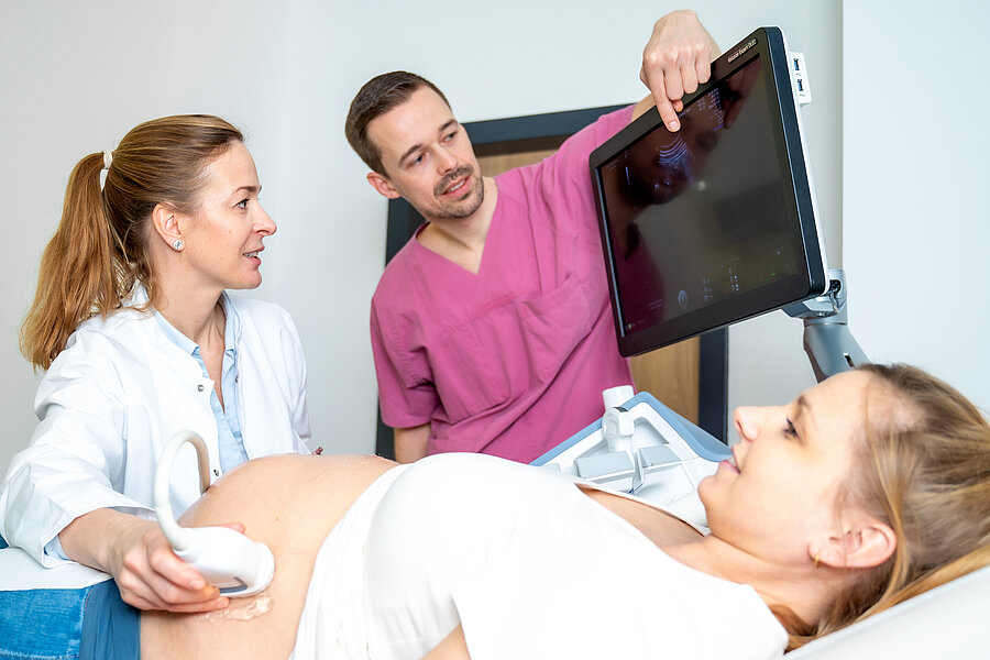 Eine Ärztin untersucht den Bauch einer schwangeren Frau mithilfe eines Ultraschallgeräts. Die schwangere Frau liegt auf einer Liege. Die Ärztin und ein weiterer Arzt schauen sich die Ultraschallbilder auf einem Monitor an. Eine Ärztin untersucht den Bauch einer schwangeren Frau mithilfe eines Ultraschallgeräts. Die schwangere Frau liegt auf einer Liege. Die Ärztin und ein weiterer Arzt schauen sich die Ultraschallbilder auf einem Monitor an.