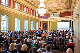 Ein Blick in den gefüllten Festsaal des Löwengebäudes am Uniplatz der Martin-Luther-Universität Halle-Wittenberg. Am weit entfernten Pult ist eine Frau zu erkennen, die spricht und dabei gestikuliert. Neben ihr wird ein historisches Bild des Löwengebäudes präsentiert.