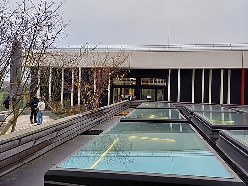 Zu sehen ist eine Dachterrasse, die auch die Oberlichter des Gebäudes beinhaltet. Diese sind wie ein Raster mit 3 mal 3 Scheiben angeordnet und schimmern türkisfarben. Das Foto wurde über die Oberlichter hinweg Richtung Gebäude aufgenommen.