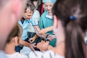 Eine Gruppe von Erwachsenen und Kindern steht um ein Übungsmodell eines menschlichen Oberkörpers, der auf einem Krankenbett liegt. Eine medizinische Fachkraft leitet einen Besucher an, eine Intubation am Modell durchzuführen. Die Aufnahme wurde zwischen den Köpfen der Besucher:innen aufgenommen, so dass deren Hinterköpfe unscharf im Vordergrund zu erkennen sind.