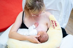 Eine Mutter stillt ihr Baby im Sitzen. Das Baby liegt auf einem mondförmigen Stillkissen. Das Kissen ist hellgelb und sieht sehr kuschelig aus. Es trägt die Aufschrift in rot: "Schön, dass du da bist". Die Mutter schaut zum Kind.