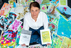 Aufnahme von oben. Eine Person sitzt zwischen vielen gemalten Bildern. Sie hält zwei Bilder in den Händen und schaut nach oben in die Kamera.