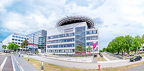 Außenansicht des Hauptgebäudes des Universitätsklinikums Halle (Saale). Vor dem Klinikum befinden sich Menschen. Am rechten und linken Bildrand stehen Bäume.