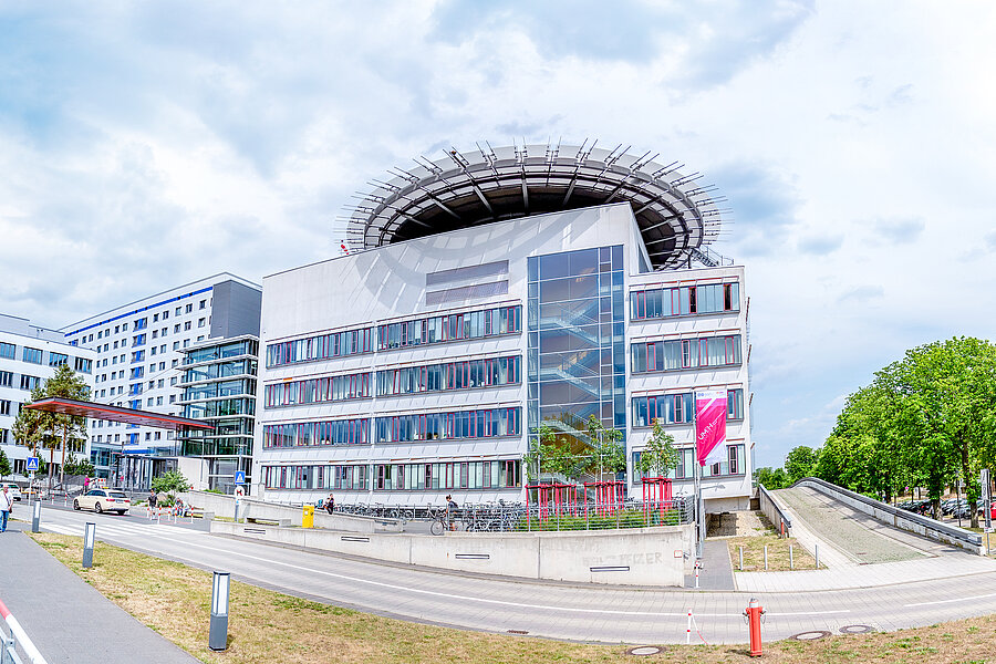 Außenansicht des Hauptgebäudes des Universitätsklinikums Halle (Saale). Vor dem Klinikum befinden sich Menschen. Am rechten und linken Bildrand stehen Bäume.  Außenansicht des Hauptgebäudes des Universitätsklinikums Halle (Saale). Vor dem Klinikum befinden sich Menschen. Am rechten und linken Bildrand stehen Bäume.