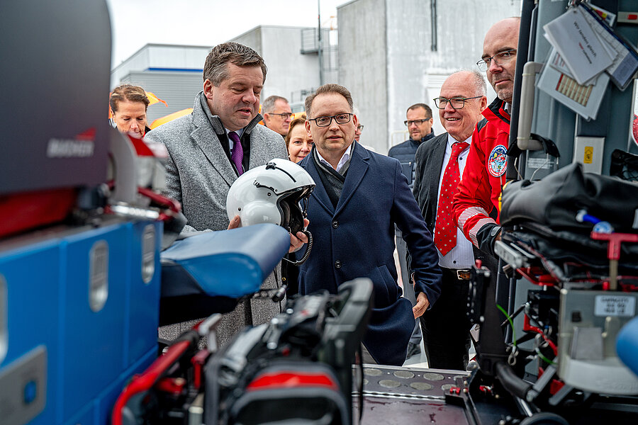 Mehrere Personen schauen in das Innere eines Rettungshubschraubers. Die Person links im Bild hält einen Helm in den Händen. Mehrere Personen schauen in das Innere eines Rettungshubschraubers. Die Person links im Bild hält einen Helm in den Händen.