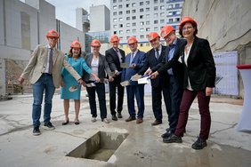 Vertreter:innen der Politik und der Universitätsmedizin Halle stehen in einer Baugrube. Alle tragen orangefarbene Helme und halten je eine Maurerkelle in ihren Händen. Die Gruppe steht im Halbkreis um ein Loch herum, in dem die Zeitkapsel versenkt wird.