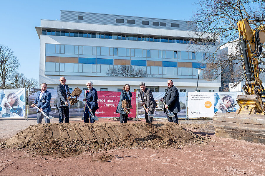 6 Personen mit Spaten an einem Erdhügel. Rechts von Ihnen ein Bagger, im Hintergrund ein Gebäude. 6 Personen mit Spaten an einem Erdhügel. Rechts von Ihnen ein Bagger, im Hintergrund ein Gebäude.