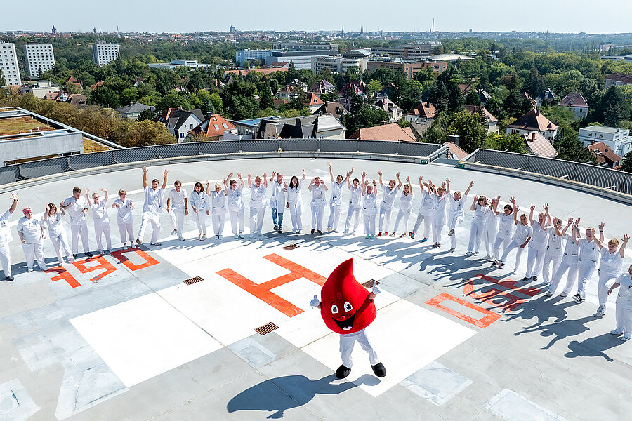 Drohnenaufnahme aus der Vogelperspektive: Eine große Gruppe von Menschen in medizinischer Arbeitskleidung steht in einem Halbkreis auf dem Hubschrauberlandeplatz des Universitätsklinikums Halle (Saale). Alle winken in die Kamera. In der Mitte des Halbkreises steht eine Person in einem Blutstropfen-Kostüm und streckt die Arme nach oben. Im Hintergrund ist ein Teil der Stadt Halle zu sehen. Drohnenaufnahme aus der Vogelperspektive: Eine große Gruppe von Menschen in medizinischer Arbeitskleidung steht in einem Halbkreis auf dem Hubschrauberlandeplatz des Universitätsklinikums Halle (Saale). Alle winken in die Kamera. In der Mitte des Halbkreises steht eine Person in einem Blutstropfen-Kostüm und streckt die Arme nach oben. Im Hintergrund ist ein Teil der Stadt Halle zu sehen.