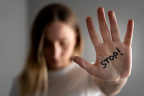Eine Frau streckt eine Hand aus, auf ihrer Handfläche steht „Stop“. Der Fokus liegt auf der Hand, ihr Kopf und Oberkörper sind unscharf.