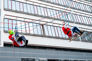 Zwei Personen, die als Grinch und als Weihnachtsmann verkleidet sind, seilen sich ab. Im Hintergrund ist ein großes Gebäude mit Fensterfront zu sehen. Der Weihnachtsmann sitzt auf einem Holzschlitten.